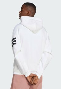 Haut à capuche blanc avec un design en V et une bande noire sur la manche gauche ; il présente une capuche avec cordon de serrage et des poignets côtelés.
