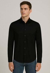 Camicia nera a maniche lunghe in tessuto liscio, con colletto a punta, bottoni frontali e fondo curvo. Indossata con jeans blu.