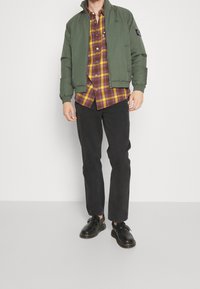 Groene bomberjas met elastische manchetten over een geruit hemd met knopen in paars, geel en bruin, gecombineerd met een zwarte broek en zwarte schoenen.