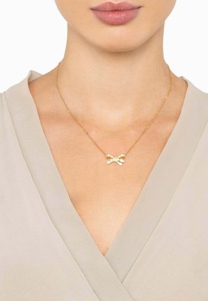 Gold necklace with a bow-shaped pendant featuring a textured design, worn on a bare neck above a light-colored, soft fabric.