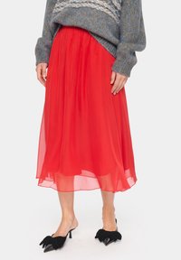 Falda roja de gasa fluida con cintura fruncida y diseño de capas. Combinada con mules negros que cuentan con destacados lazos como adorno.