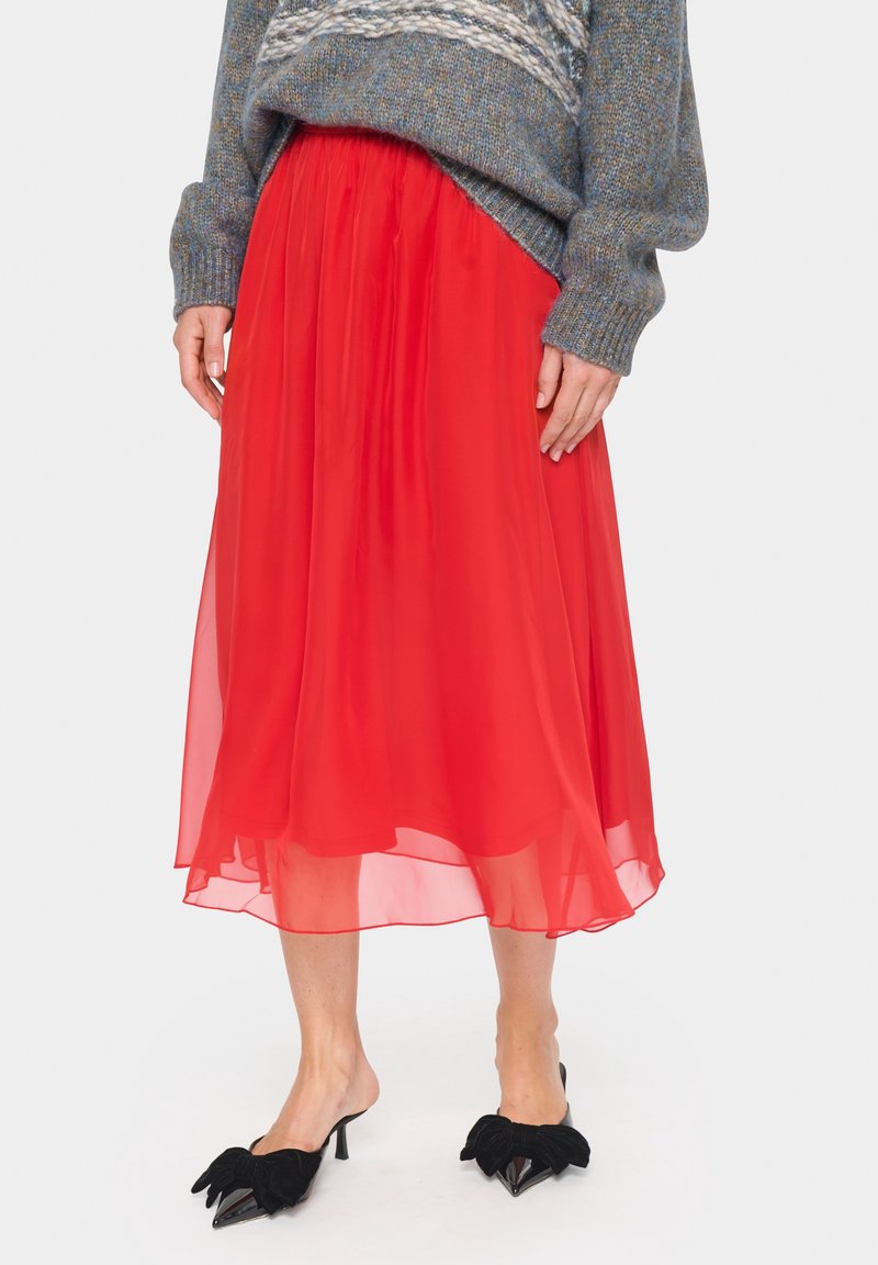 Falda roja de gasa fluida con cintura fruncida y diseño de capas. Combinada con mules negros que cuentan con destacados lazos como adorno.