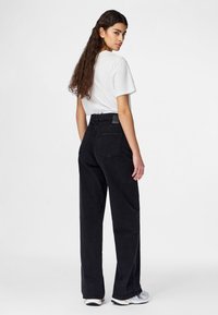 Fiatal nő hosszú sötét hajjal, fehér pólót, magas derekú fekete széles szárú farmert és fehér tornacipőt visel, és hátrafordulva néz.