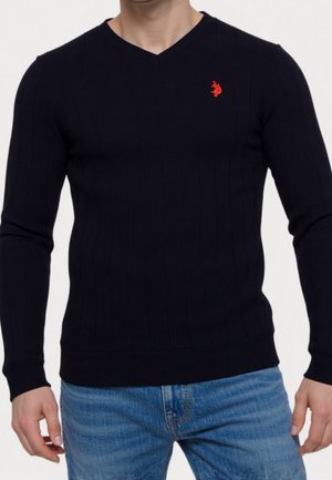 Mann trägt einen eng anliegenden schwarzen V-Ausschnittpullover mit rotem gesticktem Logo auf der Brust, kombiniert mit hellblauen Jeans.