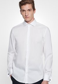 Vit långärmad skjorta med spetsig krage, framknäppning och svarta manschettakcenter, med en slät, texturerad tyg.