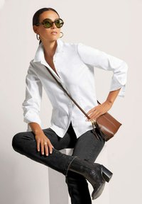 Weißes Button-up Hemd, schwarze Jeans, braune Umhängetasche, schwarze kniehohe Stiefel; runde grüne Sonnenbrille; Model sitzt mit den Händen auf den Hüften.