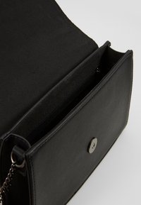 Bolso de mano de cuero negro con una textura suave, que cuenta con un compartimento con cremallera y un cierre de botón a presión. Detalle de cadena visible en la correa.