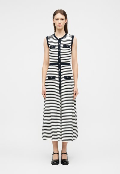 Femme portant une robe midi sans manches à rayures horizontales noires et blanches avec des boutons sur le devant et quatre poches à rabat, debout devant un fond uni.