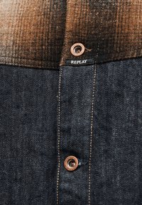 Gros plan sur un tissu à carreaux marron rencontrant un denim foncé avec des boutons en cuivre et une étiquette en tissu noire marquée « REPLAY » près de la couture.
