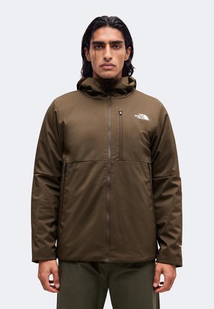 Uomo con capelli scuri che indossa una giacca North Face verde oliva con zip e cappuccio, in piedi contro uno sfondo chiaro e uniforme.