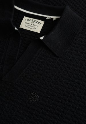 Sort tekstureret polo med et strikket mønster, der har en flad krave, hvide accenter og et broderet logo på brystet.