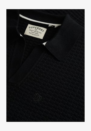Sort tekstureret polo med et strikket mønster, der har en flad krave, hvide accenter og et broderet logo på brystet.
