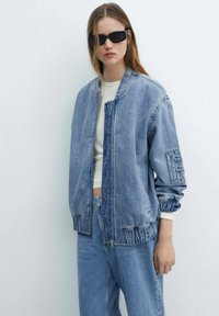 Mlada žena nosi oversized svijetloplavu traper jaknu, odgovarajuće traperice, bijeli crop top i crne pravokutne sunčane naočale na jednostavnoj bijeloj pozadini.