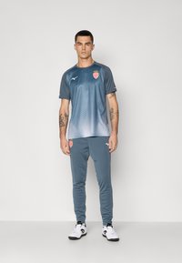 Homme adossé à un mur neutre, portant un t-shirt de sport en dégradé gris et blanc et un pantalon assorti avec des baskets blanches, tatouages visibles sur les bras.