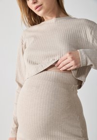 Maglietta maternità beige a coste con maniche lunghe, caratterizzata da un design corto che mette in mostra una gonna maternità a coste coordinata.