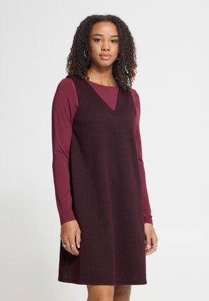 Femme aux cheveux bouclés portant un haut bordeaux à manches longues sous une robe sans manches bordeaux foncé, debout contre un fond uni clair.