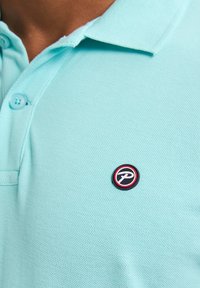 Polo shirt azzurro con tessuto texturizzato, caratterizzato da un colletto a due bottoni e una toppa rotonda con logo nei toni del rosso e nero sul petto.