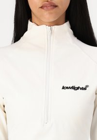 Bijela zip-up flis jakna s visokim ovratnikom, s crnim izvezenim logom. Glatka tekstura s vidljivim detaljima šivanja, bez uzoraka.