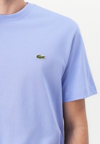 Blaues Baumwoll-T-Shirt mit rundem Ausschnitt. Auf der Brust befindet sich ein gesticktes grünes Krokodil-Logo, es hat kurze Ärmel und eine glatte Textur.