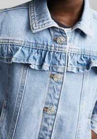 Ljusblå denimmantel med volangkrage, silverfärgad knäppning och vertikala sömdetaljer. Mjuk, texturerad tyg.