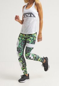 Sportovní souprava obsahující bílý síťovaný top s černou grafikou a elastické legíny v zeleném a černém maskáči s bílými akcenty.