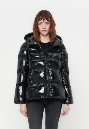 Donna che indossa una lucida giacca a vento nera con cappuccio, chiusura con zip e tasche frontali, abbinata a jeans grigio scuro.