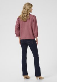 Blusa rosa con maniche a sbuffo e righe verticali abbinata a jeans a zampa d'elefante blu scuro e scarpe col tacco color beige. Sfondo uniforme.