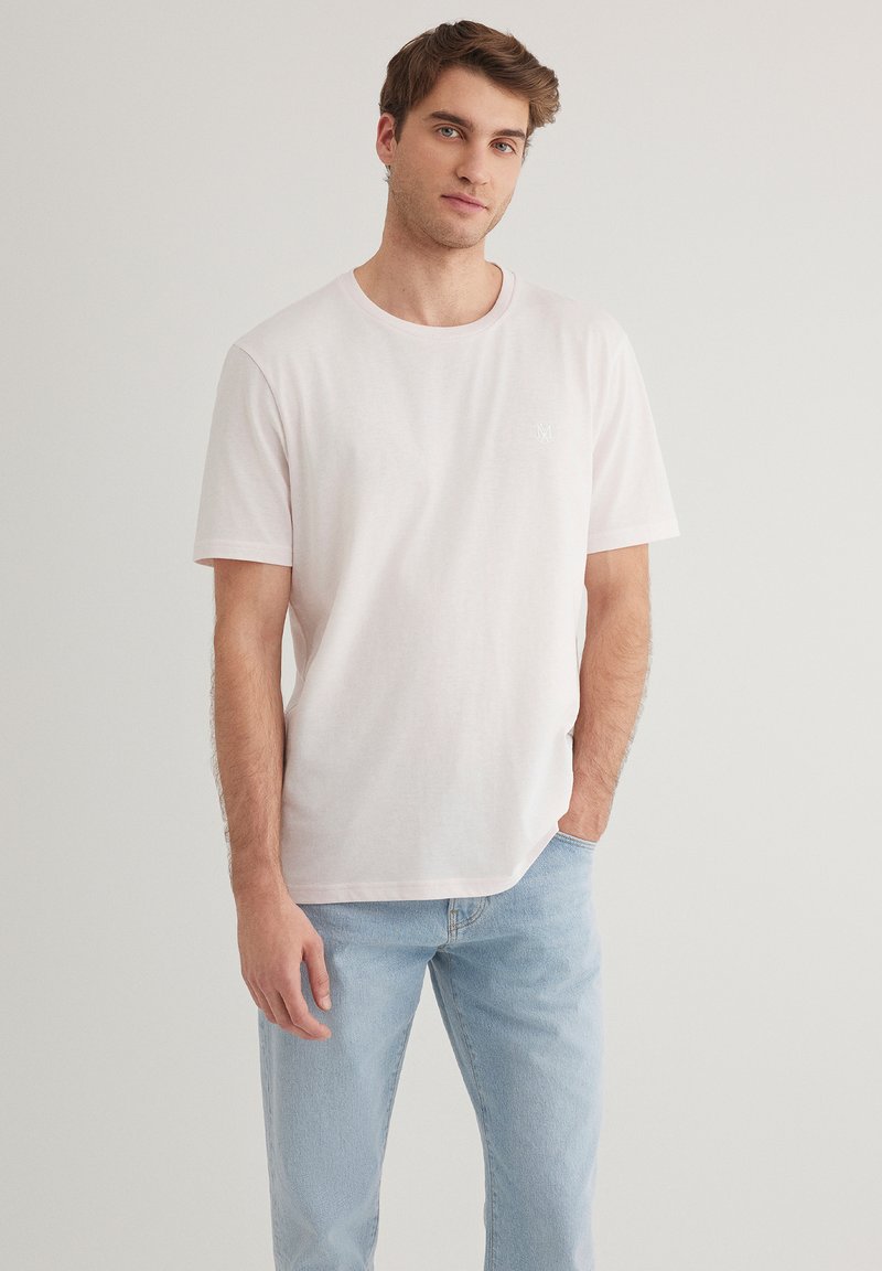 Helles rosa Baumwoll-T-Shirt mit kurzen Ärmeln, Rundhalsausschnitt und einem kleinen Logodetail auf der Brust. Kombiniert mit hellblauen Jeans.