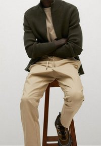 Chaqueta acanalada verde oliva con cuello alto, combinada con pantalones beige claros que tienen puños elásticos. Calzado marrón con cordones y detalles.
