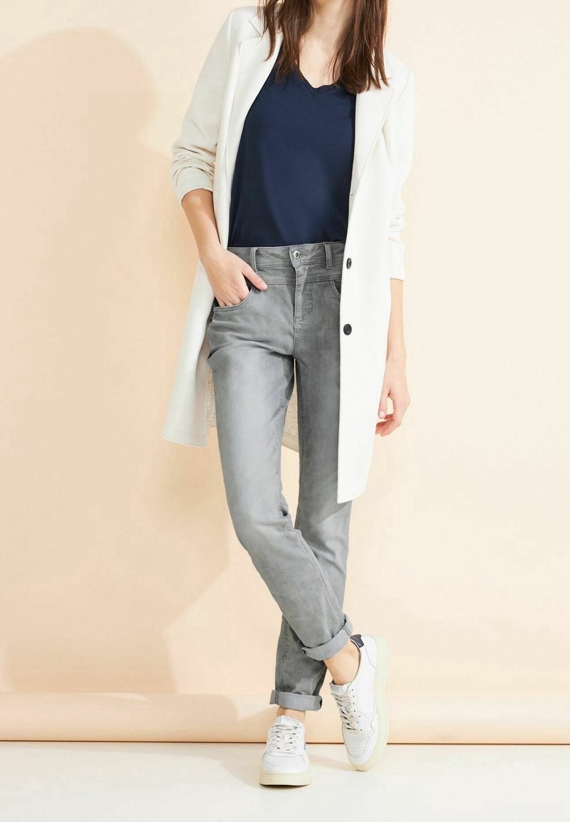 Femme portant un long blazer blanc, un top bleu marine, un jean gris retroussé et des baskets blanches, debout avec une main dans la poche.