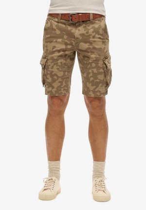 Mand iført beige camouflage cargoshorts, brunt læderbælte, beige sokker og lysebeige sneakers står med fødderne let adskilt.