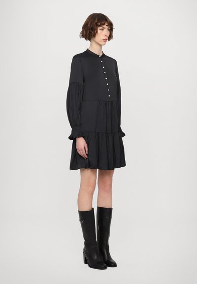 Zwarte jurk met lange geplooide mouwen, een knoopsluiting aan de voorkant en een gelaagde rok. Gestyled met zwarte laarzen met hoge hakken tot de knie.