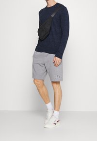 Námořnický pletený svetr, šedé fleecové šortky s logem, černá křížová taška, bílé tenisky s červenými a modrými akcenty. Neformální outfit.