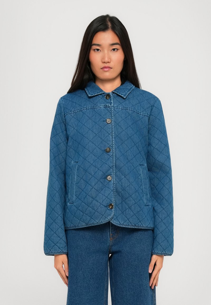Femeie îmbrăcată într-o jachetă albastră matlasată, închisă la butoane, cu pantaloni din denim albastri asortati, stând pe un fundal deschis simplu.