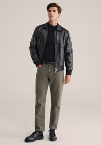 Jachetă bomber din piele ecologică neagră, cu fermoar frontal, mansete cu dantelă, purtată peste un tricou negru, asortată cu pantaloni verzi măsliniu și pantofi negri.