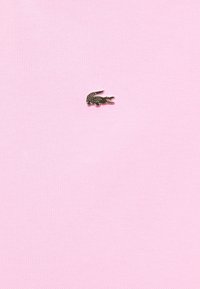 Helle rosa Strukturstoff mit einem kleinen metallischen, dunkel gefärbten Krokodil-Logo auf der unteren Seite.