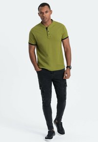 Polo shirt verde oliva con dettagli neri, dotato di colletto con bottoni. Abbinato a pantaloni cargo neri e sneakers nere. Design semplice e aderente.
