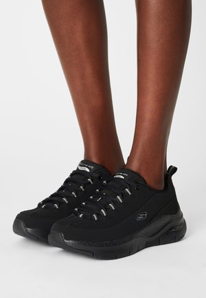 Jambes à la peau foncée portant des baskets à lacets Skechers noires avec des semelles épaisses, posées sur une surface blanche unie.
