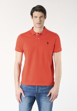 Uomo che indossa una polo arancione a maniche corte con piccolo logo nero sul petto e jeans blu, in piedi davanti a uno sfondo bianco semplice.