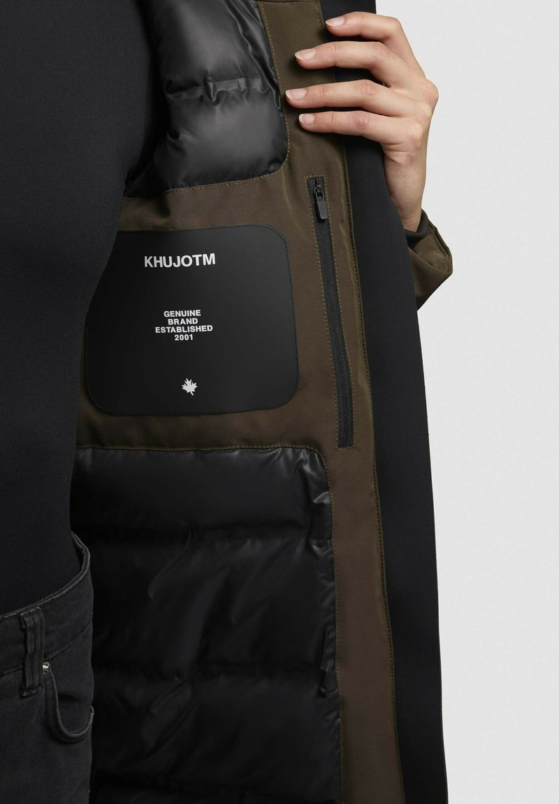 Interior de chaqueta acolchada con forro acolchado negro y tela verde oliva. Presenta un bolsillo con cremallera y una etiqueta con el nombre de la marca y el año de establecimiento.