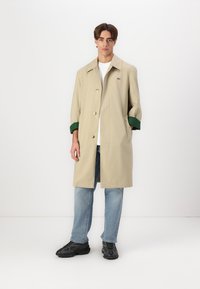 Beige vandtæt frakke med en klassisk pasform, der har grønne manchetter, knapper og sidelommer. Kombinationen med lyseblå jeans og sorte sneakers.