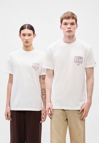 Valkoiset lyhythihaiset T-paidat, joissa pieni, värikäs graafinen logo rinnassa. Molemmilla malleilla on rento, neutraalin sävyn housut.