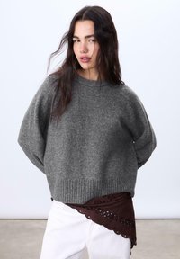 Grauer, gestrickter Pullover mit weit geschnittenen Ärmeln und geripptem Saum, getragen über einem gemusterten Stoff in Weinrot. Das Outfit wird mit weißen Hosen kombiniert.