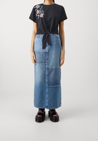 Zwarte top met bloemrijke borduurwerk, geknoopt aan de voorkant. Lange denim rok met patchworkdesign en rafelige randen. Zwarte plateau schoenen.