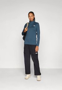 Dolga majica s teal rokavi, z visokim ovratnikom in logotipom, združena s črnimi cargo hlačami z žepi. Model nosi bež superge in ima na sebi nahrbtnik.