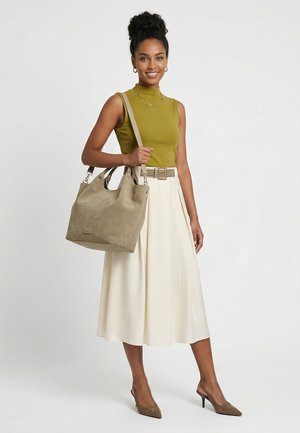 Frau trägt olivgrünes ärmelloses Top und cremefarbenen Midirock, trägt große beigefarbene Handtasche, lächelt und posiert vor einfarbigem Hintergrund.