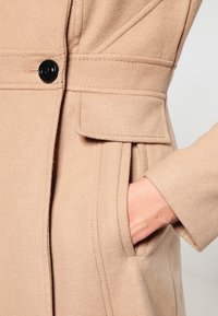 Abrigo de lana beige con un solo botón negro, bolsillo lateral y detalles de costura a lo largo de las costuras para aportar textura.