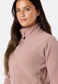 Jasnoróżowa polarowa kurtka zapinana na zamek z wysokim kołnierzem. Posiada haftowane logo, gładką teksturę oraz detale na bocznych szwach. Doskonała do noszenia na co dzień.