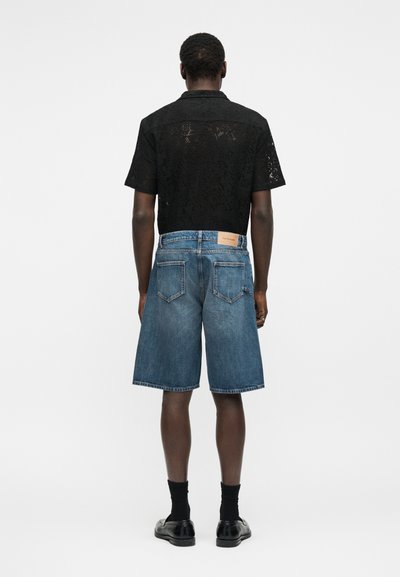 Άνδρας που φοράει ημιδιαφανές μαύρο πουκάμισο με κοντά μανίκια, μπλε τζιν σορτς μέχρι το γόνατο, μαύρες κάλτσες και μαύρα παπούτσια, στέκεται γυρισμένος με την πλάτη.