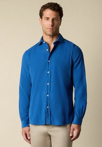 Man in een lichtblauwe overhemden met lange mouwen en beige broek, staand tegen een effen beige achtergrond.